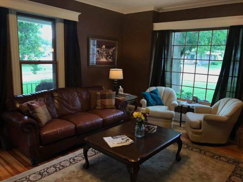 Dark brown room with wood floor, brown leather couch, two beige chairs, a coffee table, and large windows