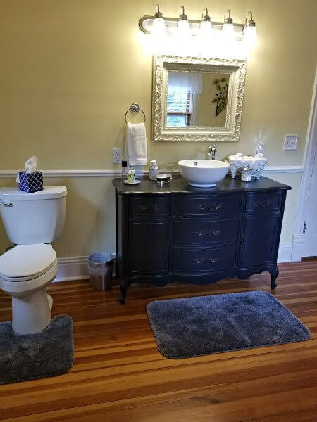 Yellow and white bathroom with wood floor, dark stained ornate vanity with white vessel sink, and blue rugs