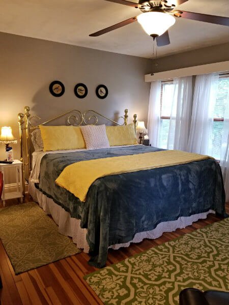 Grey room with wood floor, brass bed with blue and yellow blankets and flanking nightstands and lamps with fringed shades