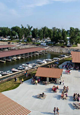 Public concrete area with red-roofed shelters and picnic tables, next to open and covered boat docks