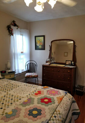 Beige room with quilt-covered bed, window with white sheers, wood dresser with mirror and small ladder back chair